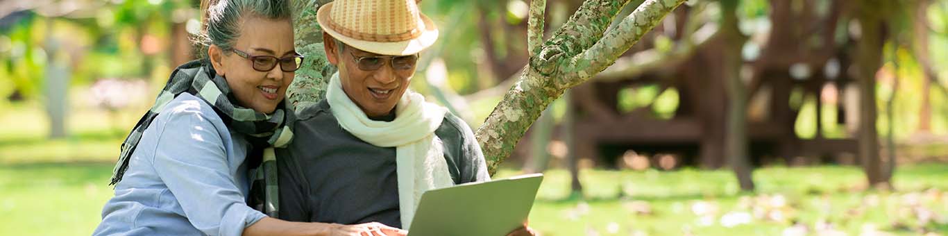 senior couple sitting on a bench at a park, wearing scarves, smiling, and reading something on a laptop screen
