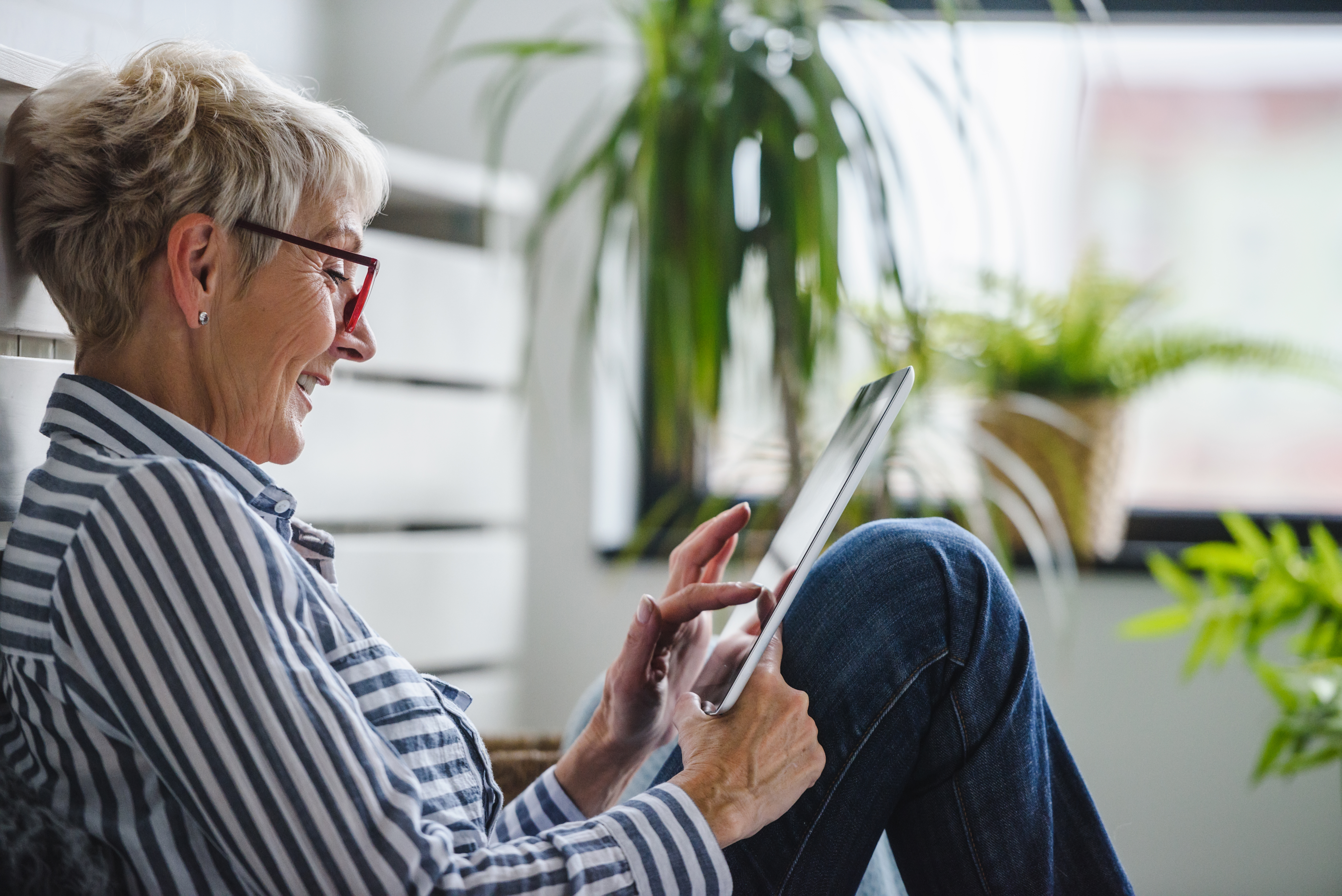 Older woman sitting in a bright room next to a houseplant, smiling and interacting with a tablet in her lap